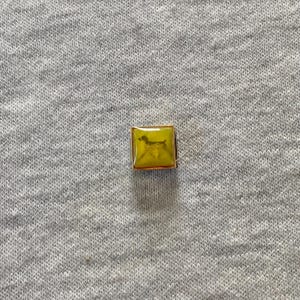 Può includere: Un ciondolo quadrato con una cornice in metallo dorato e un inserto giallo lucido. Il centro dell'inserto giallo presenta una piccola silhouette scura di un cane in piedi di profilo. Il ciondolo ha una superficie piatta e liscia.