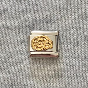 以下が含まれることがあります： 磨かれたシルバー調の金属ベースと、渦を巻いた羊の頭の形をしたゴールド調の金属製エンブレムが浮き彫りになった長方形のモジュラーチャームリンク。リンクは滑らかな金属仕上げで、水平なデザイン構造が特徴です。