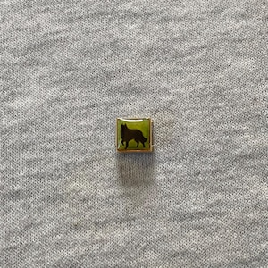 May include: A small, square charm with a gold-coloured frame. The charm features a silhouette of a dog in dark brown against a green background. It is likely a piece of jewellery or a decorative accessory.