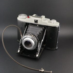 May include: A vintage Kodak 66 folding camera with a black leatherette body, a grey top plate, and a black bellows. The camera features a silver lens assembly with visible aperture markings and is attached to a long, thin black cable shutter release.
