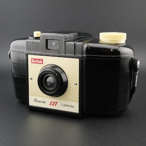 May include: A black Kodak Brownie 127 camera featuring a cream-coloured front plate with a gold grid pattern. The camera includes a central lens, a top-mounted cream-coloured film advance knob, a small shutter button, and the text Kodak Brownie 127.