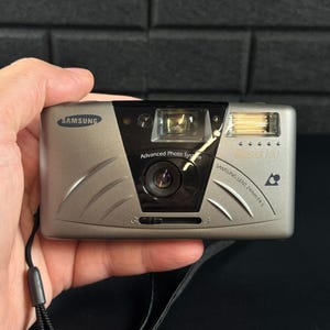 May include: A silver Samsung Fino 100 compact camera featuring an Advanced Photo System lens, a 24mm f/4.5 lens, a built-in flash, and a black wrist strap. The camera body has a metallic finish with curved decorative lines on the front panel.