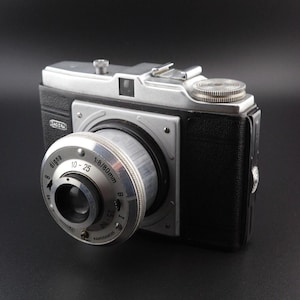 May include: A vintage Digna camera with a black textured body and silver metal top plate. The lens barrel features the text 'digna', '1:8/80mm', '10-25', 'B', 'I', and 'KAMERAWERK'. It includes a silver shutter release button and a film advance knob.