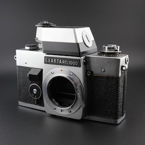 May include: A silver and black Exakta RTL1000 35mm SLR camera body. The camera features a prominent central lens mount, a textured black grip, a top-mounted prism viewfinder, and various mechanical dials and controls on the top plate.