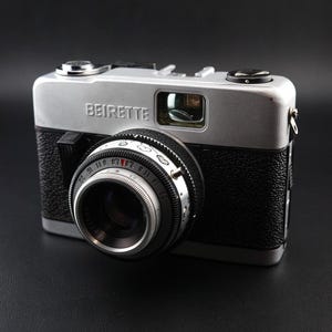 May include: A vintage Beirette film camera featuring a silver top plate and a black textured body. The lens barrel displays aperture settings, shutter speeds, and a series of small heart icons. The word BEIRETTE is printed on the front panel.
