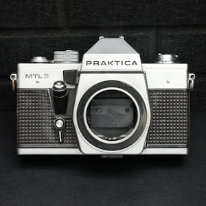 May include: A silver and black Praktica MTL3 camera. The camera has the word "PRAKTICA" printed on the top and "MTL3" on the side. The camera has a textured grip and a large lens opening.