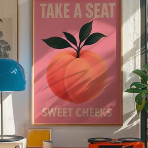 Puede incluir: Un p&oacute;ster rosa enmarcado con el texto "TAKE A SEAT" y "SWEET CHEEKS" encima y debajo de una ilustraci&oacute;n de melocot&oacute;n. El p&oacute;ster est&aacute; en un marco dorado y se exhibe en una habitaci&oacute;n con un tocadiscos y otra decoraci&oacute;n.