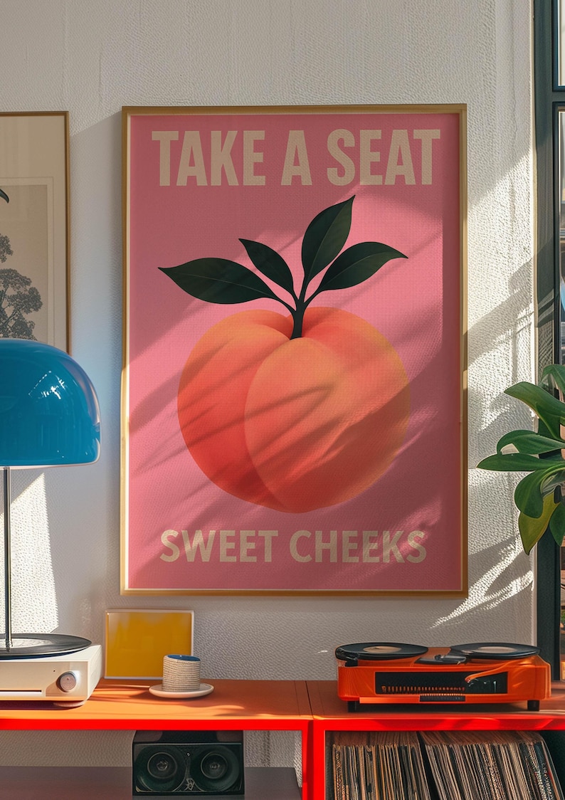 Puede incluir: Un p&oacute;ster rosa enmarcado con el texto "TAKE A SEAT" y "SWEET CHEEKS" encima y debajo de una ilustraci&oacute;n de melocot&oacute;n. El p&oacute;ster est&aacute; en un marco dorado y se exhibe en una habitaci&oacute;n con un tocadiscos y otra decoraci&oacute;n.