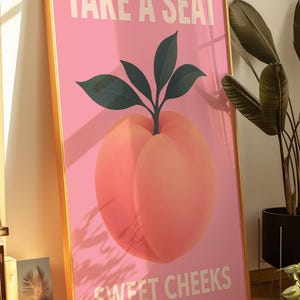 Puede incluir: Un estampado art&iacute;stico rosa enmarcado con una ilustraci&oacute;n de melocot&oacute;n y el texto "TAKE A SEAT SWEET CHEEKS" en blanco. El melocot&oacute;n tiene hojas verdes y un marco dorado. La obra de arte est&aacute; apoyada contra una pared.