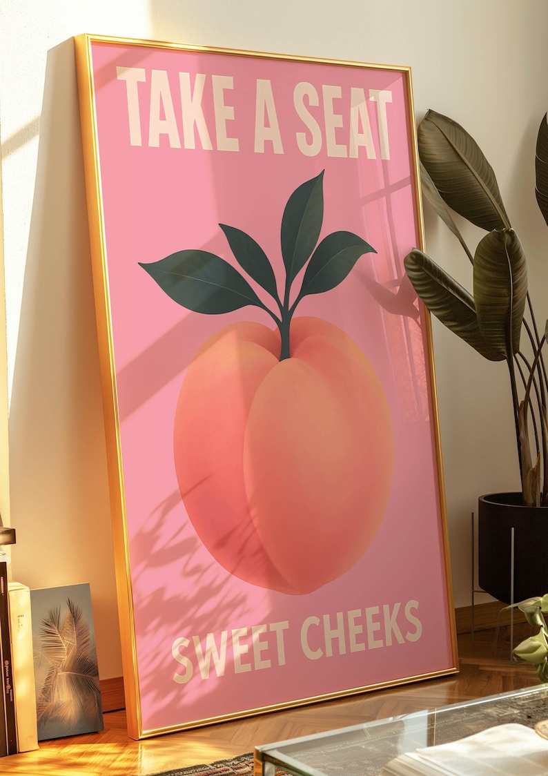 Puede incluir: Un estampado art&iacute;stico rosa enmarcado con una ilustraci&oacute;n de melocot&oacute;n y el texto "TAKE A SEAT SWEET CHEEKS" en blanco. El melocot&oacute;n tiene hojas verdes y un marco dorado. La obra de arte est&aacute; apoyada contra una pared.