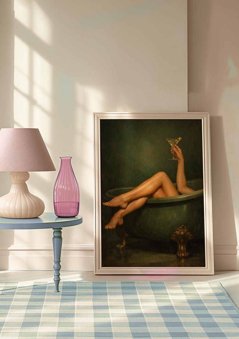 May include: A framed artwork depicts a person relaxing in a green bathtub, holding a cocktail. The artwork is propped against a wall. A pink lamp and vase sit on a blue side table, with a blue and white checkered rug in the foreground.