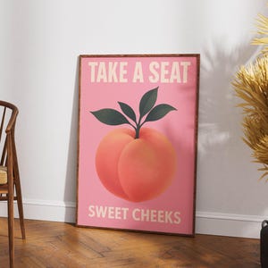 Puede incluir: Un p&oacute;ster rosa enmarcado con una ilustraci&oacute;n de melocot&oacute;n y el texto "TAKE A SEAT SWEET CHEEKS" en blanco. El melocot&oacute;n es de color naranja claro con hojas verdes. El marco es marr&oacute;n.