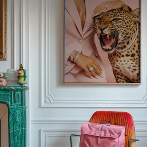 May include: A framed artwork depicts a woman in a pink suit with a bejeweled leopard. A pink satchel and a smaller pink handbag sit on a colorful chair. The room features a green fireplace and white walls.