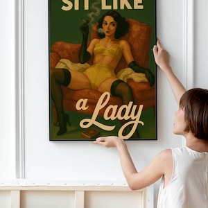May include: A framed art print featuring a vintage-style illustration. The artwork depicts a woman in yellow lingerie, black gloves, and stockings, seated in a red armchair, smoking a cigarette. The text "SIT LIKE a Lady" is displayed.