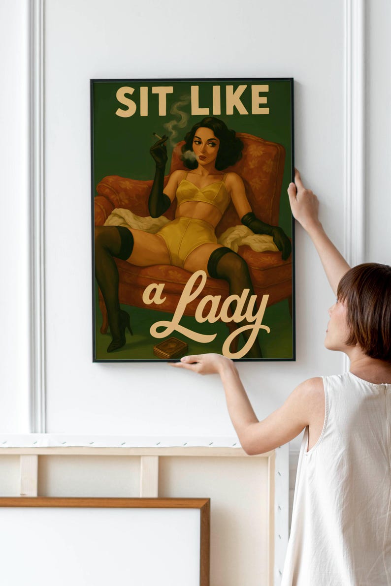May include: A framed art print featuring a vintage-style illustration. The artwork depicts a woman in yellow lingerie, black gloves, and stockings, seated in a red armchair, smoking a cigarette. The text "SIT LIKE a Lady" is displayed.