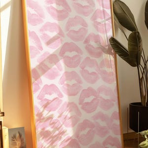 May include: A framed artwork featuring a pattern of pink lip prints on a white background. The frame is gold-colored, and the artwork is leaning against a wall. The image is well-lit, with a plant in the background.
