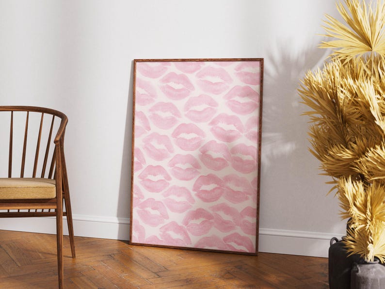 May include: A framed art print featuring a pattern of pink lips on a white background. The artwork is displayed in a room with a wooden floor and a wooden chair. The print has a brown frame.