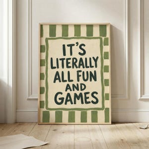 May include: A rectangular wall art print featuring a cream-colored center with the dark green hand-lettered text IT'S LITERALLY ALL FUN AND GAMES, surrounded by a thick dark green border and a wide outer frame of alternating green and cream stripes.
