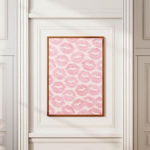 May include: A framed print featuring a pattern of pink lips on a white background. The artwork is displayed in a room with white trim and a wooden frame. The lips are in a repeating pattern.