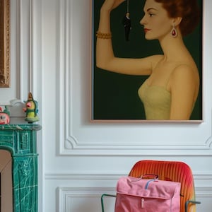 以下が含まれることがあります： 額縁に入った絵画には、紐で小さな男性を抱える女性が描かれています。女性は黄色のドレスと赤いイヤリングをしています。ピンクのブリーフケースと小さなピンクのバッグがカラフルな椅子の上にあります。部屋には白い壁と緑の暖炉があります。