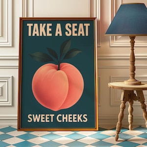 Puede incluir: Impresión artística enmarcada con una ilustración de melocotón. La obra presenta un melocotón grande y maduro en tonos naranja y rosa sobre un fondo verde azulado. El texto "TAKE A SEAT" y "SWEET CHEEKS" se muestra en blanco.