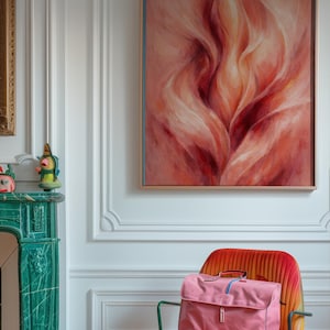 May include: An abstract painting in a wooden frame hangs above a vibrant orange chair. A pink satchel bag with a blue stripe sits on the chair. The room features a green fireplace and white walls.
