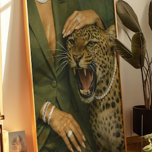 May include: Framed artwork featuring a leopard with an open mouth, adorned with a diamond necklace. The artwork includes a person wearing a green blazer and diamond jewelry. The frame is gold.