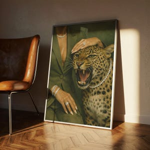 May include: A framed art print featuring a leopard with an open mouth, wearing a diamond necklace, and a person in a green suit. The person is wearing diamond jewelry. The artwork is displayed next to a brown leather chair.