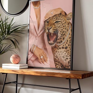 May include: Framed art print featuring a leopard with an open mouth, wearing a diamond necklace. A person in a pink blazer and jewelry is touching the leopard. The art is displayed on a wooden bench.