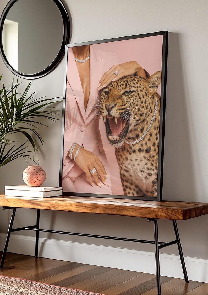 May include: Framed art print featuring a leopard with an open mouth, wearing a diamond necklace. A person in a pink blazer and jewelry is touching the leopard. The art is displayed on a wooden bench.