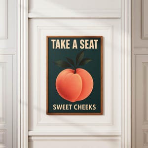 May include: A framed art print with a peach illustration and the text "TAKE A SEAT" and "SWEET CHEEKS". The peach is orange with green leaves, set against a dark teal background. The frame is a warm brown color.