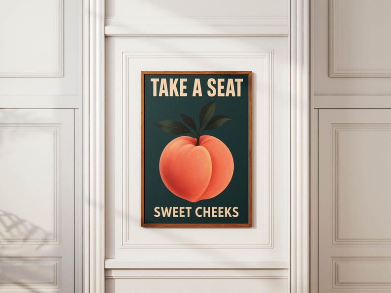 May include: A framed art print with a peach illustration and the text "TAKE A SEAT" and "SWEET CHEEKS". The peach is orange with green leaves, set against a dark teal background. The frame is a warm brown color.