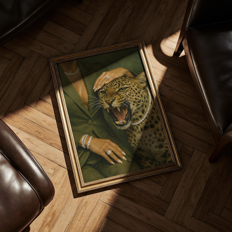 May include: A framed artwork featuring a leopard with an open mouth, wearing a pearl necklace, and being touched by a person wearing a green blazer and jewelry. The frame is gold-colored.