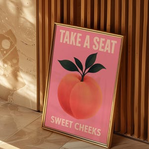 Puede incluir: Impresi&oacute;n art&iacute;stica enmarcada con fondo rosa, con una ilustraci&oacute;n de melocot&oacute;n y el texto "TAKE A SEAT SWEET CHEEKS" en blanco. El melocot&oacute;n es de un naranja-rosa vibrante con hojas verdes. El marco es dorado.