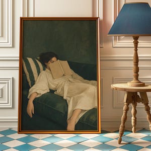 May include: A framed artwork depicts a person resting on a green sofa with a book covering their face. The person wears a cream-colored robe. A striped pillow and a wooden lamp with a blue shade are visible.