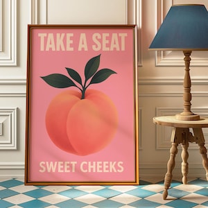 Puede incluir: Impresión artística enmarcada con una ilustración de melocotón sobre fondo rosa. El texto "TAKE A SEAT" y "SWEET CHEEKS" en beige claro. El marco es dorado. Una pieza decorativa para el hogar.
