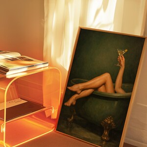 May include: A framed artwork depicts a person in a green bathtub holding a martini. The artwork is leaning against a wall next to an orange acrylic side table with books and a black and white patterned rug.