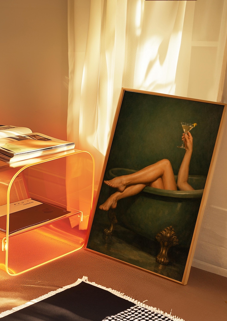 May include: A framed artwork depicts a person in a green bathtub holding a martini. The artwork is leaning against a wall next to an orange acrylic side table with books and a black and white patterned rug.