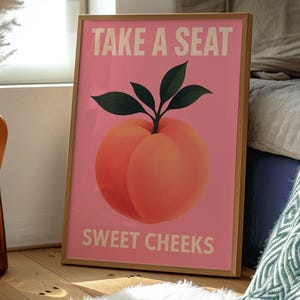 Puede incluir: Una impresi&oacute;n art&iacute;stica enmarcada con un fondo rosa presenta una ilustraci&oacute;n de melocot&oacute;n y el texto "TAKE A SEAT SWEET CHEEKS" en blanco. El melocot&oacute;n tiene un degradado de naranja y rojo con hojas verdes.