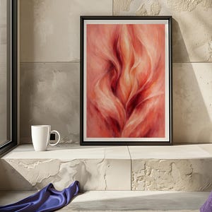 May include: Framed abstract art print with a black border. The artwork depicts a flowing, flame-like design in red and peach hues. A white mug is on a ledge beside the framed art. A blue fabric and patterned rug are also visible.