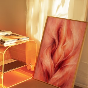 May include: A framed abstract painting in shades of red and orange. The artwork depicts flowing, organic forms, suggesting motion. A clear acrylic side table with books and a black and white patterned rug are also visible in the scene.
