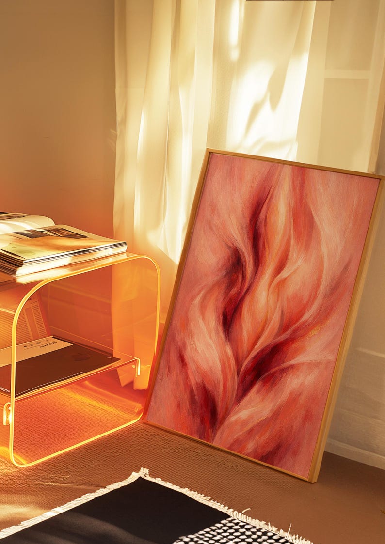May include: A framed abstract painting in shades of red and orange. The artwork depicts flowing, organic forms, suggesting motion. A clear acrylic side table with books and a black and white patterned rug are also visible in the scene.