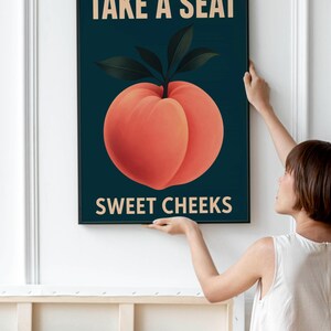 May include: A framed art print with a peach illustration and dark green leaves on a dark teal background. The text "TAKE A SEAT" and "SWEET CHEEKS" are in a light beige colour.