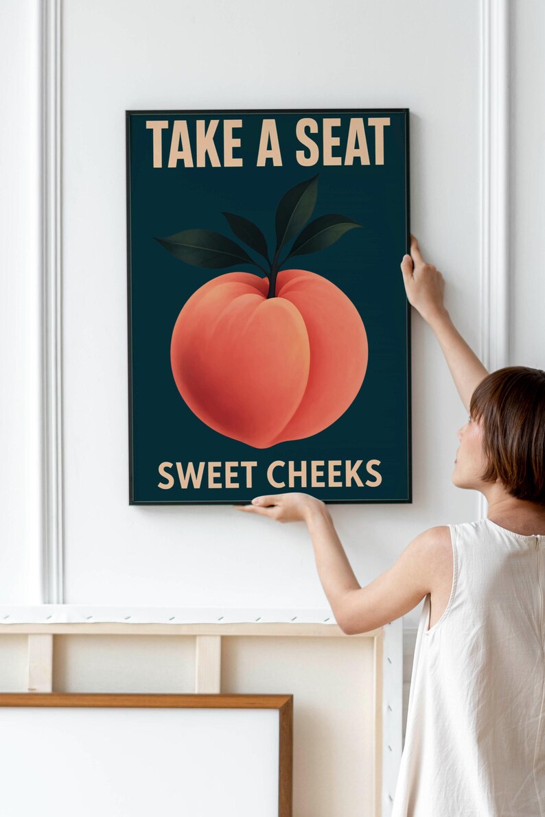 May include: A framed art print with a peach illustration and dark green leaves on a dark teal background. The text "TAKE A SEAT" and "SWEET CHEEKS" are in a light beige colour.