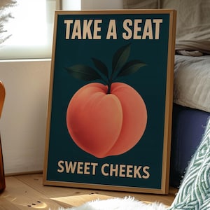 May include: A framed art print featuring a peach illustration with the text "TAKE A SEAT" above and "SWEET CHEEKS" below. The peach is a vibrant orange-red colour with green leaves, set against a dark teal background. The frame is a light brown colour.