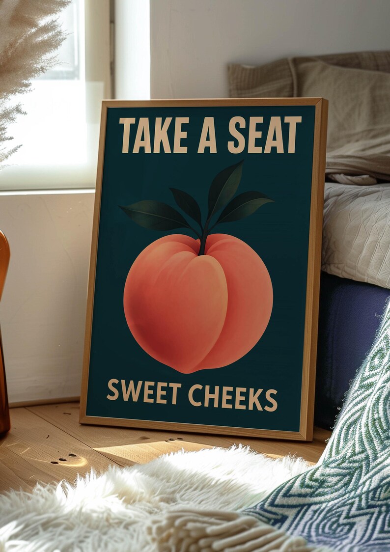 May include: A framed art print featuring a peach illustration with the text "TAKE A SEAT" above and "SWEET CHEEKS" below. The peach is a vibrant orange-red colour with green leaves, set against a dark teal background. The frame is a light brown colour.