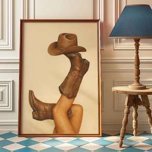 May include: Framed artwork depicting brown cowboy boots and a matching cowboy hat. The boots have detailed decorative stitching. The artwork is presented in a gold-coloured frame.