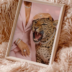 May include: Framed artwork featuring a leopard wearing a diamond necklace, with a woman in a pink suit and jewelry. The leopard's mouth is open, and the artwork is set against a pink background and a fluffy, peach-colored surface.