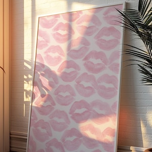May include: A framed art print featuring a pattern of pink lipstick kiss marks on a white background. The artwork is leaning against a white brick wall, with sunlight casting shadows across the surface. The frame is white.