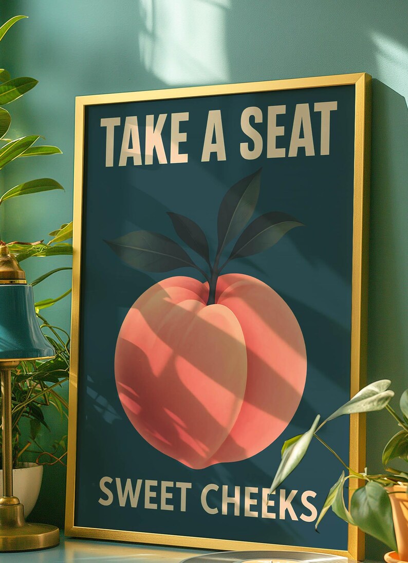May include: Framed art print with a gold border. The artwork features a large peach illustration with the text "TAKE A SEAT SWEET CHEEKS" in gold lettering against a dark teal background.